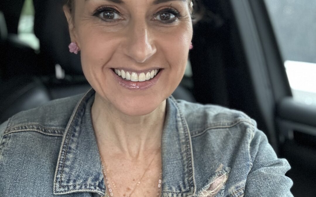 Jeanette smiling brightly wearing a denim jacket and light pink cotton shirt, pink earrings. She is in the car, which is where she spends much of her time running errands.